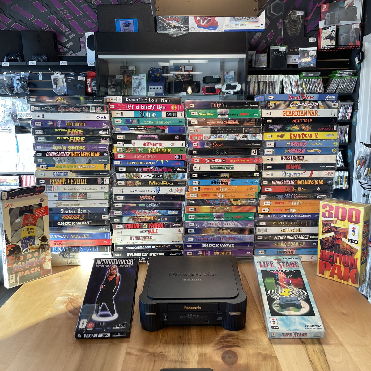 A Panasonic 3DO REAL console on a wood counter, flanked by stacked 3DO game cases including Night Trap and the 3DO Action Pak